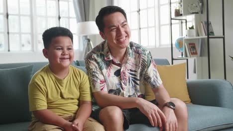 happy asian father and his son are watching tv while spending time together at home