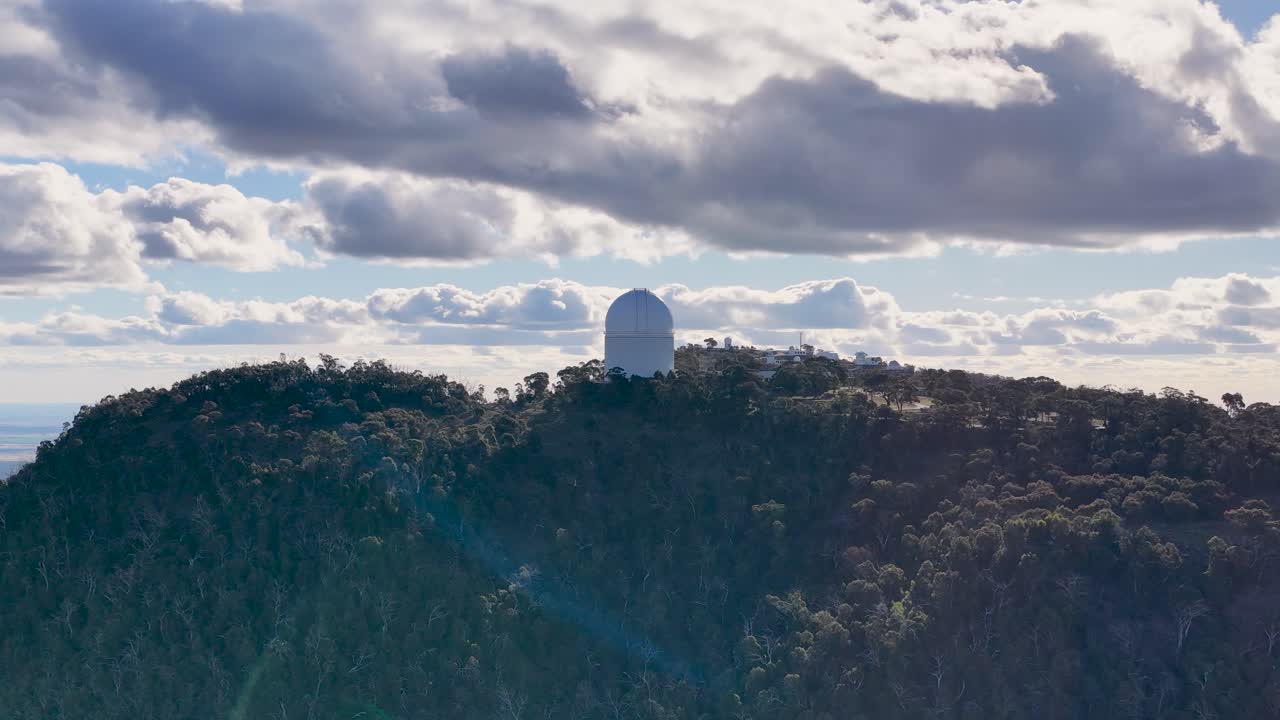 Drone captures a smooth panoramic view of a white observatory dome atop a wooded mountain under dramatic, cloud-filled skies with soft daylight