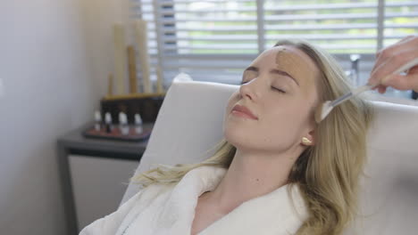 young blonde caucasian woman receive a health skin care facial treatment while lying on a massage bed in luxury resort spa saloon