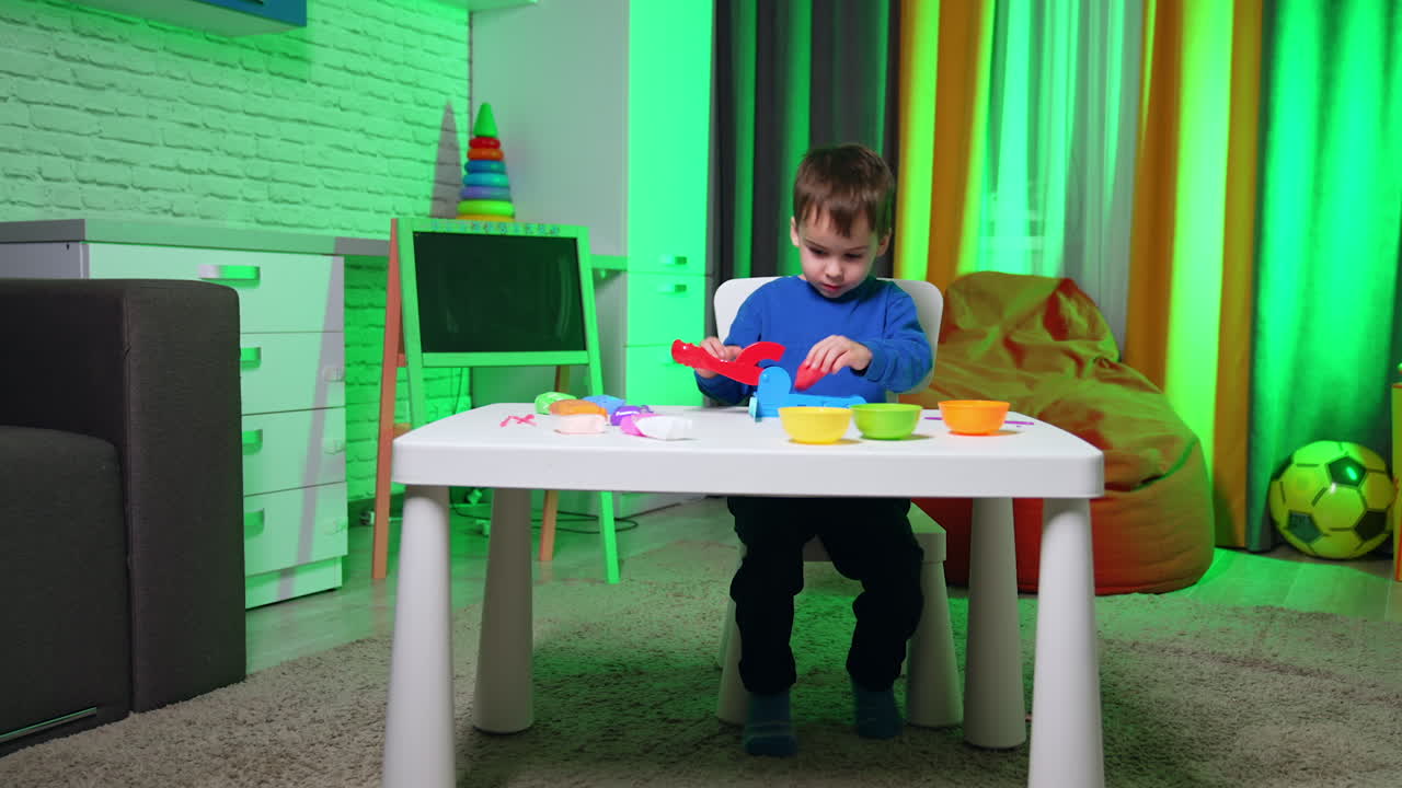 Lovely toddler kid sits at white desk at home. Focused baby boy plays with plasticine and mold machine.
