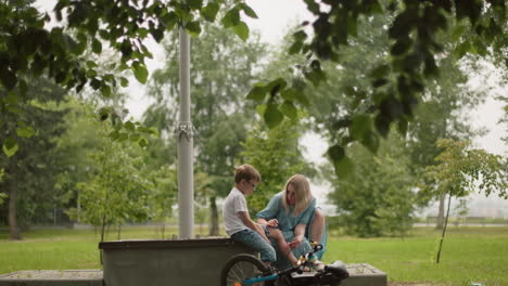 a mother gently attends to her son s wound while seated beside him on a bench in a park, surrounded by greenery, the boy holds his trousers as his mother carefully cleans his leg injury