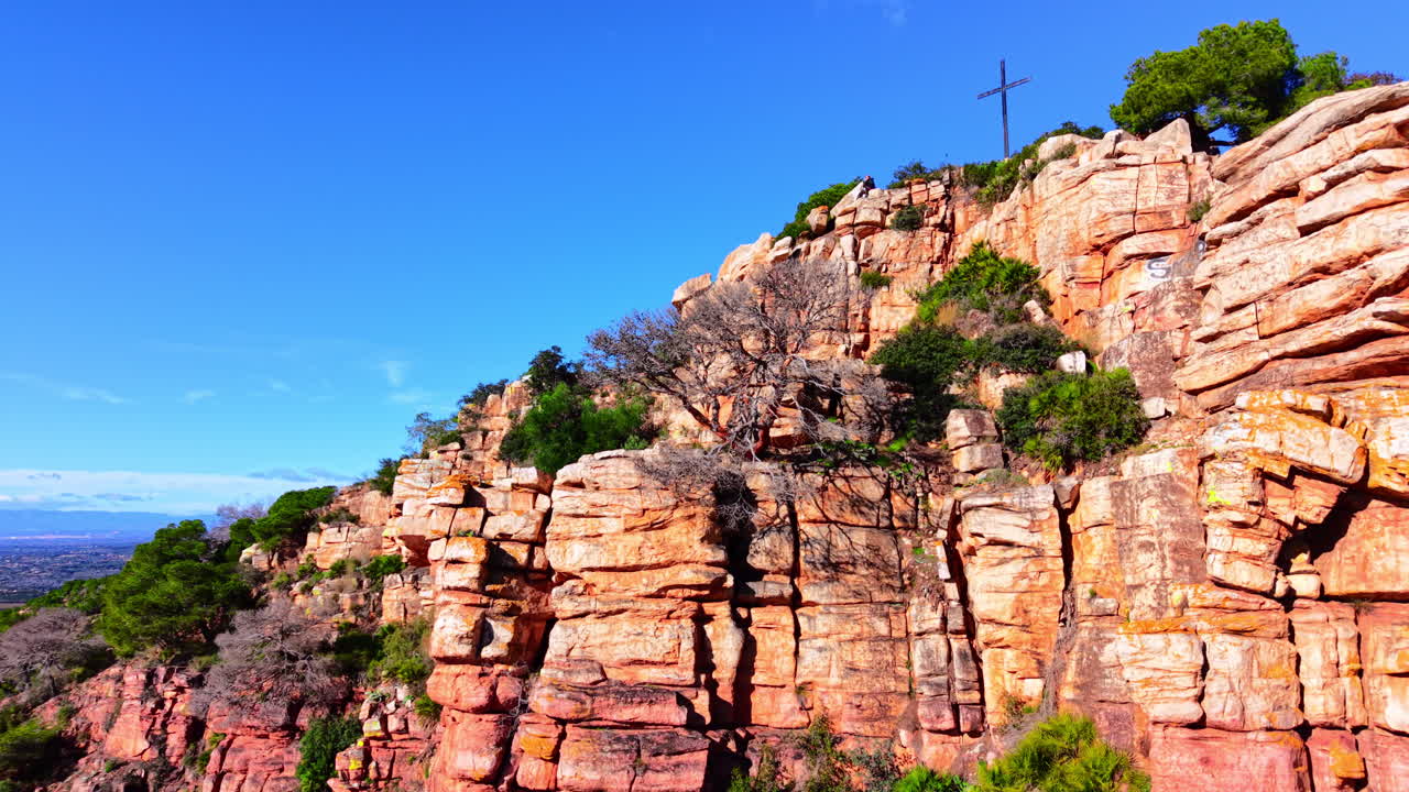 Scogliere rosse stratificate con vegetazione rada e una croce in cima sotto un cielo azzurro limpido. Formazioni rocciose di arenaria rossa con pini. Vista aerea.