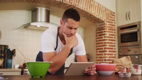hombre caucásico con un delantal sonriendo mientras usa una tableta digital en la cocina en casa