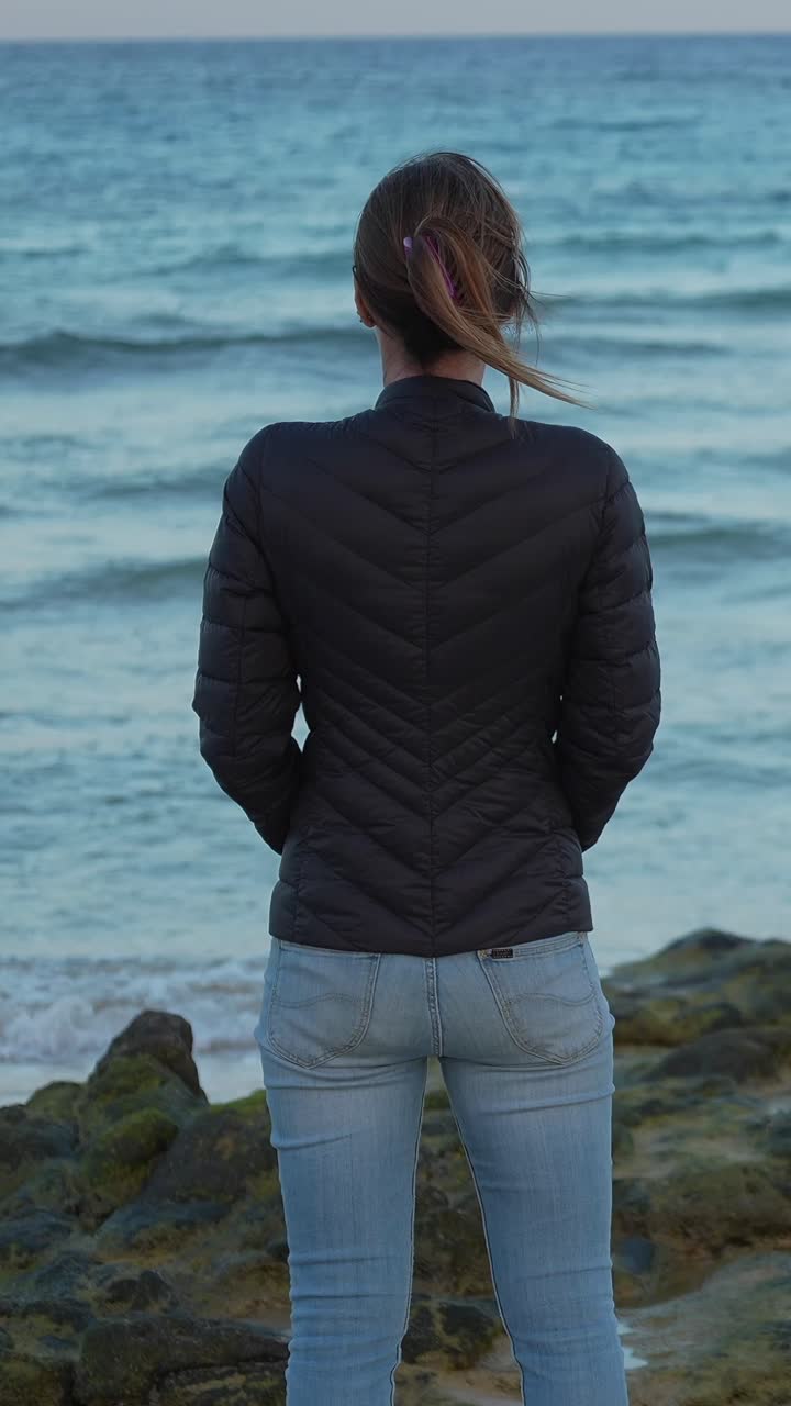 Solitary woman standing on a rocky beach, looking at the calm sea during an autumn day
