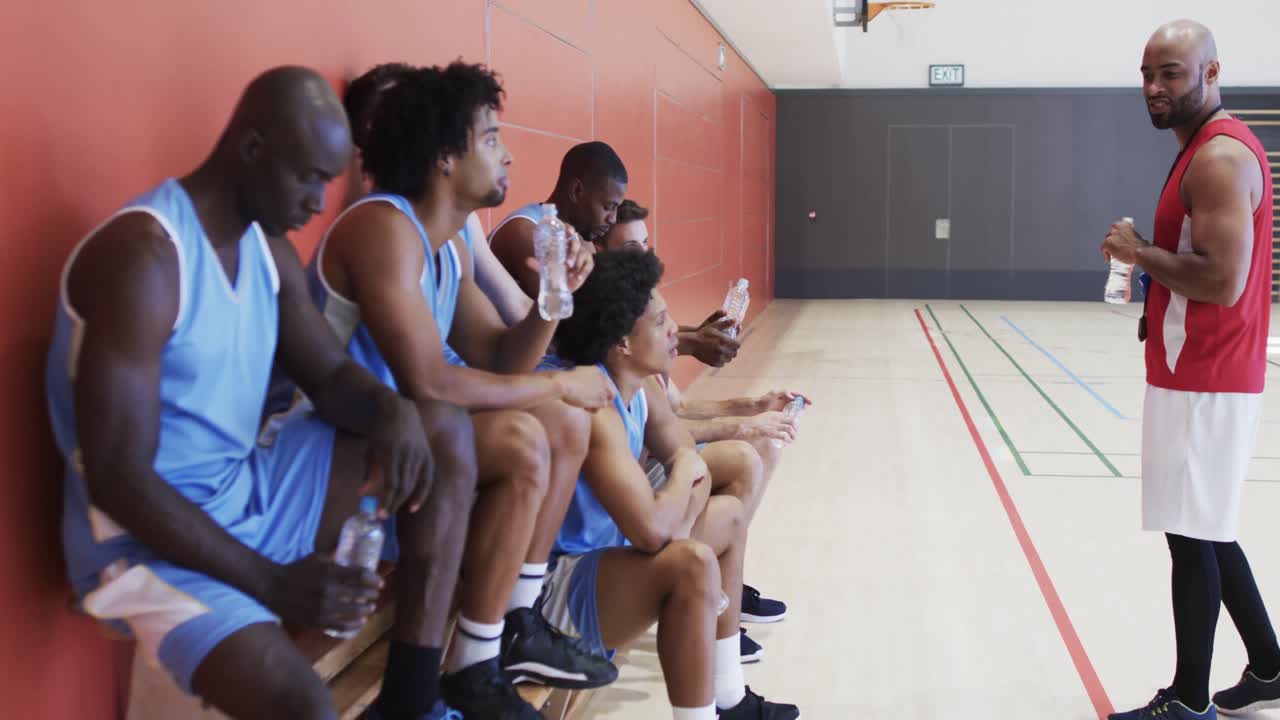entrenador de baloncesto masculino diverso hablando con el equipo sentado en el banco en la cancha cubierta, cámara lenta