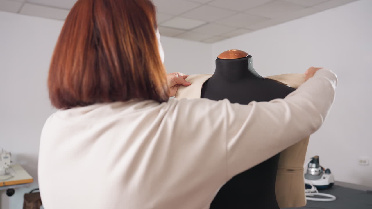 Cheerful seamstresses in fashion studio as one carefully places completed beige jacket onto black mannequin, admiring design outcome while adjusting garment shoulders with attention to detail