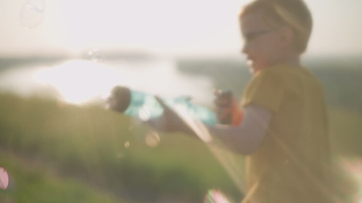 Young boy wearing a yellow shirt and glasses, smiling and running under bright sunlight, holding a plastic bubble gun that releases bubbles. moment with a strong sun flare effect