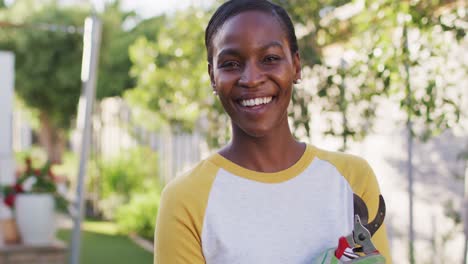 Portrait-of-african-amercian-woman-holding-secateur-looking-to-camera-and-smiling-in-garden