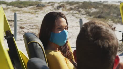 Happy-caucasian-couple-wearing-face-masks-sitting-in-beach-buggy