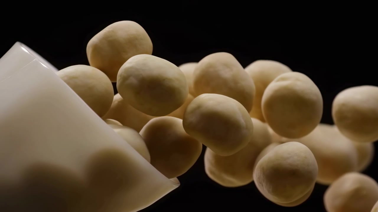 Une vue rapprochée capture des pièces rondes et pâles se déplaçant d'un récipient blanc sur une surface sombre, avec un éclairage chaud mettant en valeur des textures lisses, des formes regroupées et de subtils changements de position pendant le mouvement