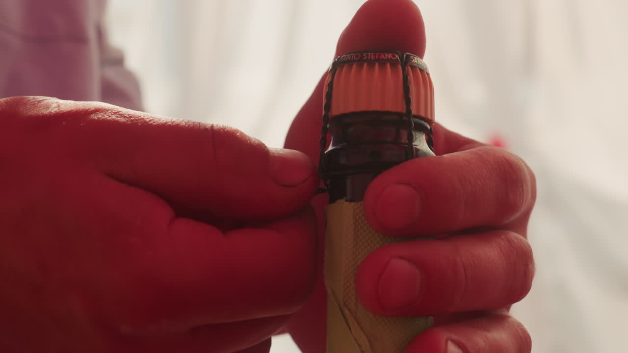 primo piano di mani che tolgono la gabbia dallo champagne, dita che stringono il filo di ferro e la stagnola, tensione che sale prima che il tappo salti. L'illuminazione calda e lo sfondo drappeggiato creano un'atmosfera intima e romantica in cui i ruoli passano da host