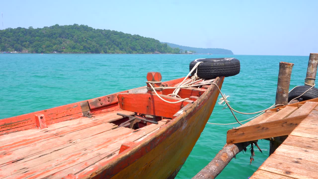barco de madera atado al muelle, balanceándose suavemente con las olas, isla tropical en segundo plano