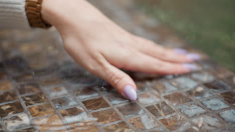 poised feminine hand with polished nails wearing watch gently skimming water surface of textured mosaic fountain, soft flowing jets and ripples half visible in blurred background