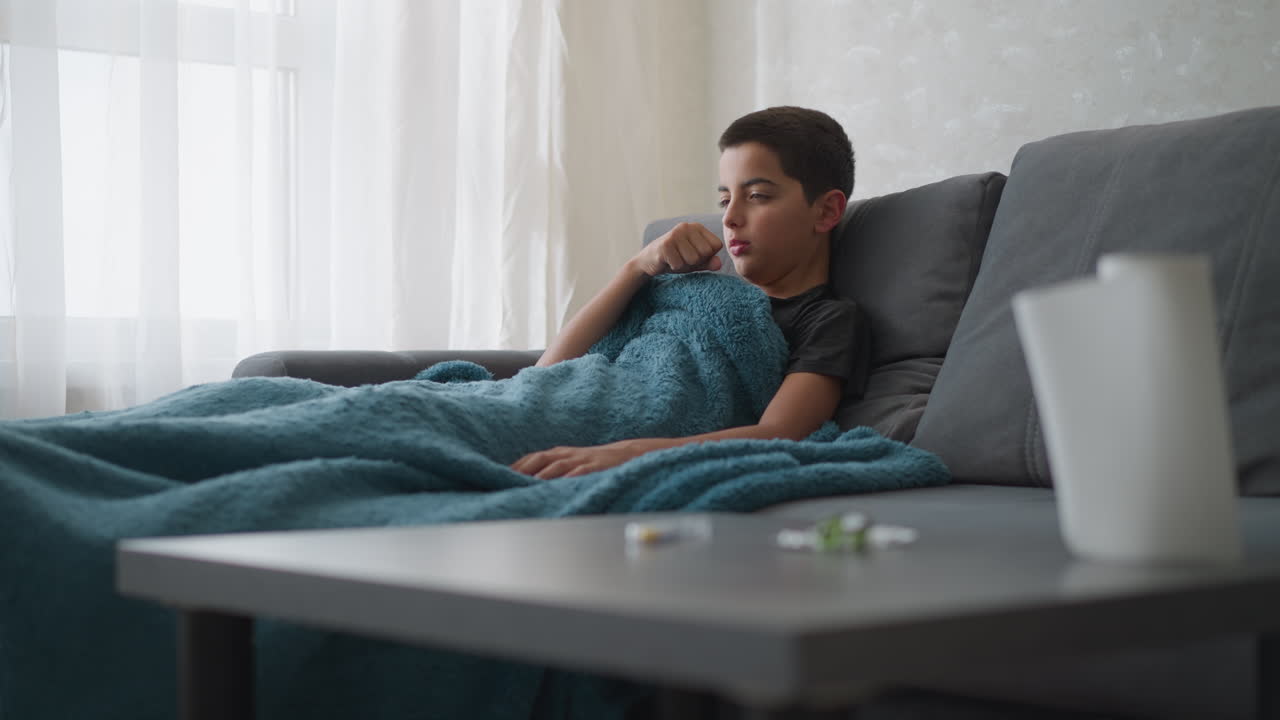 Bambino piccolo con tosse influenzale sdraiato sul divano, coperto da una spessa coperta, mentre si riposa vicino alla finestra con la tenda, con medicine e fazzoletti sfocati visibili sul tavolo in primo piano