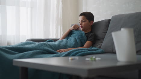 young child suffering from flu cough lies on couch with thick blanket covering him, as he rests near window with curtain, with blurred medicine and tissue visible on table in foreground