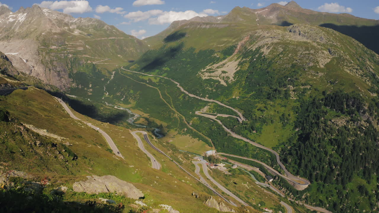 Panorama of green alpine valley with winding serpentine road carved on steep slopes under a mostly sunny sky. Furka Pass, Switzerland. Tripod, pan right, warm.
