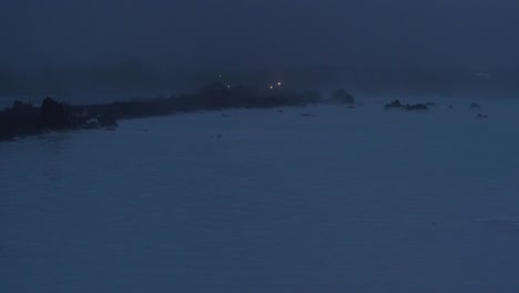 Island,-Blaue-Lagune,-Geothermisches-Kraftwerk-Svartsengi-Bei-Nacht,-Entfernte-Gestalt,-Die-Auf-Einem-Pfad-über-Dem-Wasser-Spazieren-Geht,-Weitwinkelaufnahme