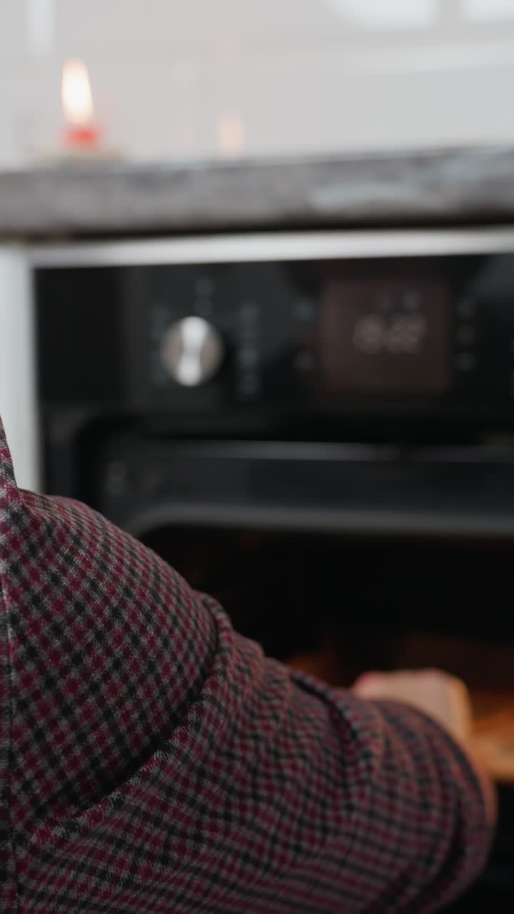 Donna vista da dietro che controlla i biscotti nel forno, chiudendo lo sportello dopo aver ispezionato, candela rossa accesa sul ripiano della cucina moderna con un'atmosfera calda e accogliente