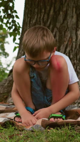 un niño pequeño se sienta bajo un árbol, profundamente pensativo, con un moretón notable en la pierna, sus manos apoyadas en el suelo, y una manta debajo de él