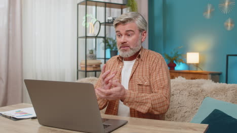 mature man sitting at table with laptop making serious business video call conversation at home