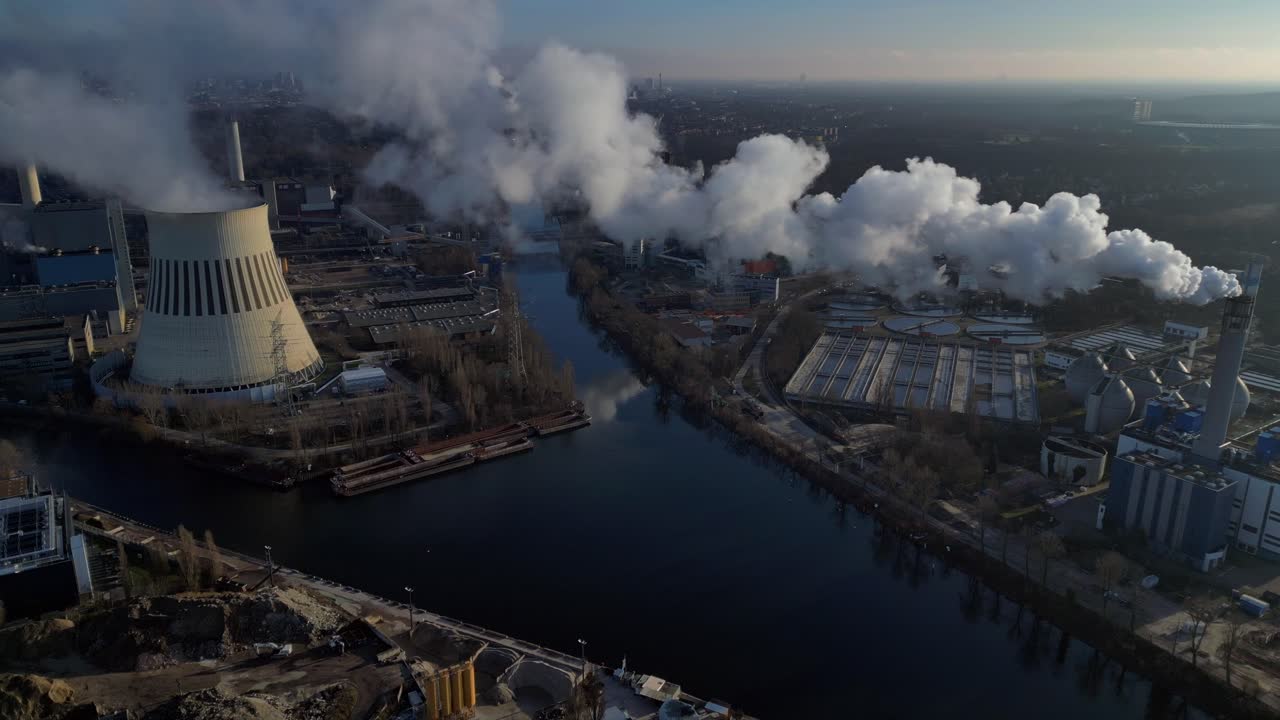 large industrial power plant emitting large amounts of smoke over the city Berlin next to a river on a sunny day. Best aerial view flight static tripod hovering drone