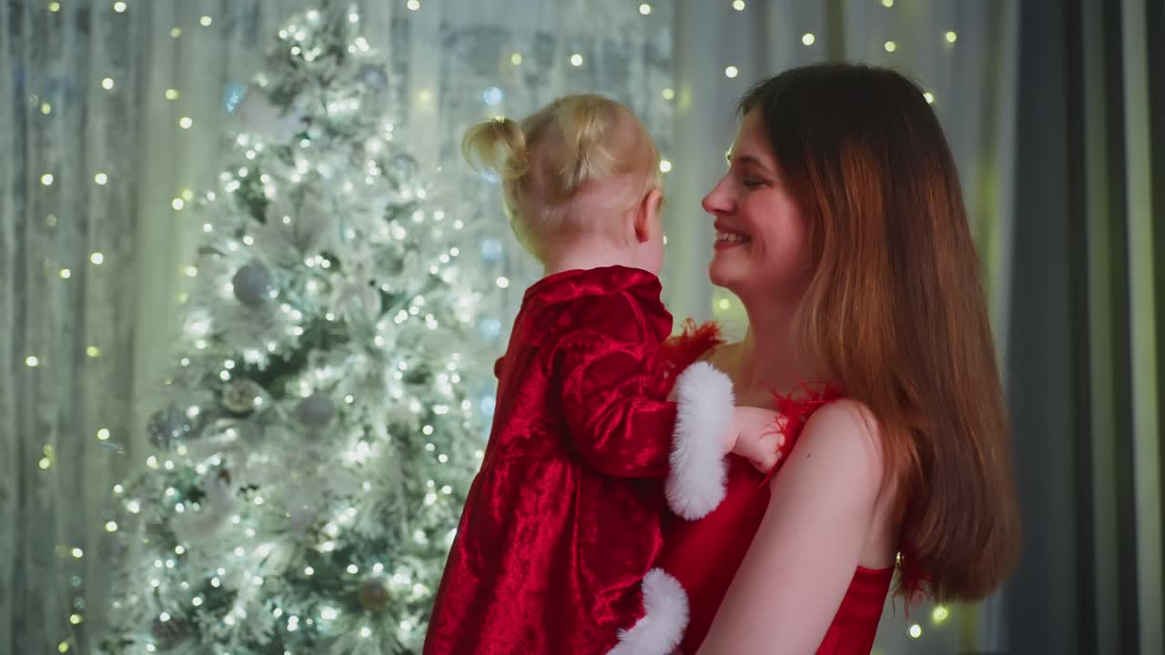 Loving mother cuddling daughter in red velvet Christmas dress, sharing tender moments amid warmly lit festive home decor