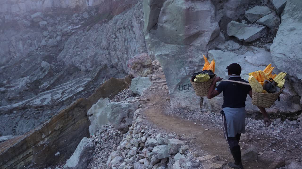 Handheld static shot shows a lone miner trekking a narrow cliff path out of Kawah Ijen crater, shouldering two woven baskets packed with bright yellow sulfur slabs