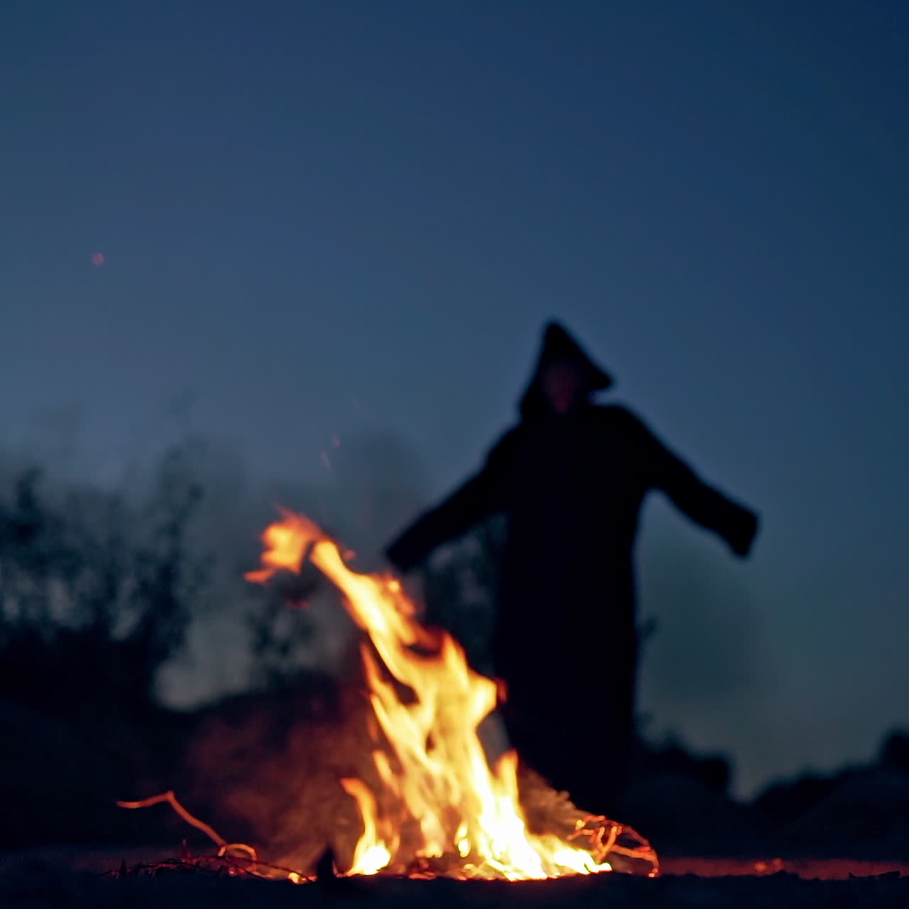 Witch at night outdoors. Flame from fire on the ground and silhouette of evil walking nearby and making fear. Halloween night.