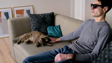 side view of a blind man in sunglasses sitting on the sofa at home and petting his cat 1