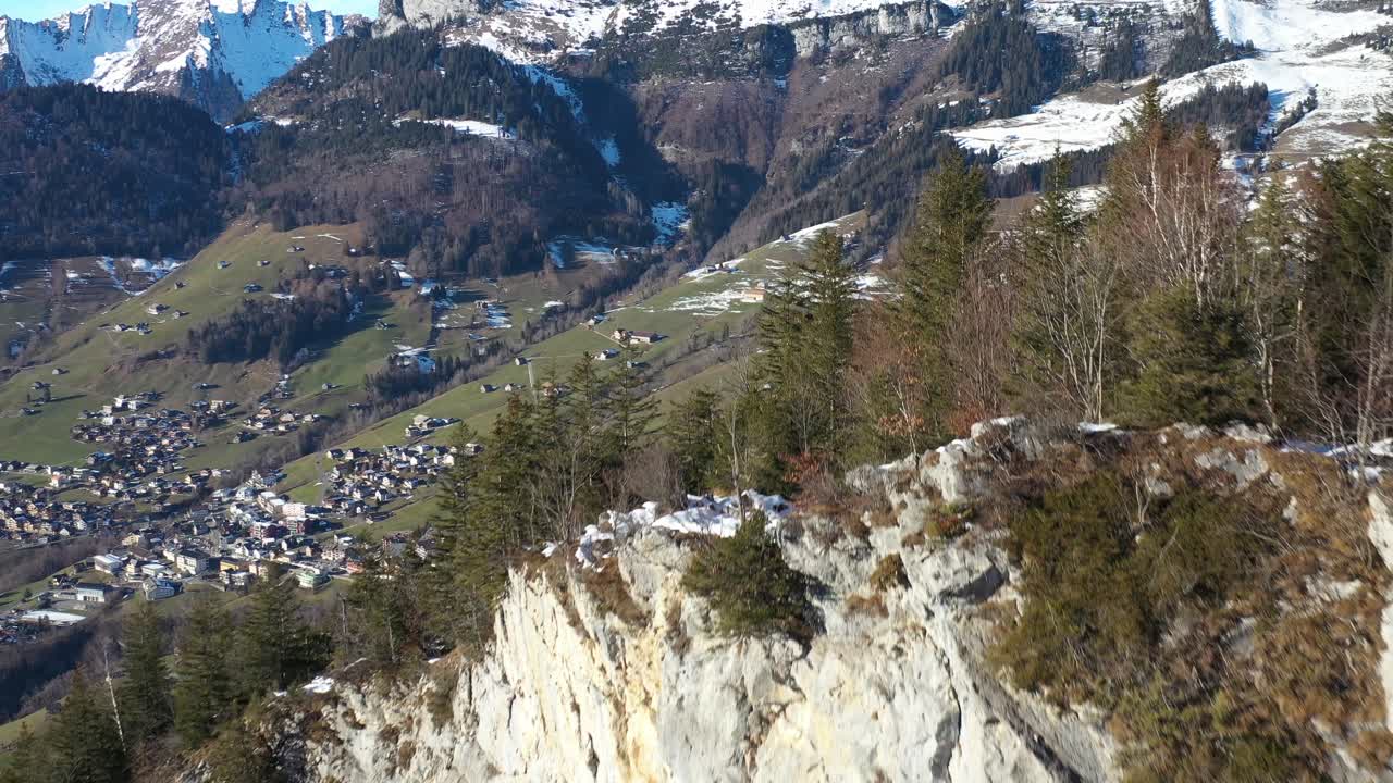 sobrevuelo aéreo de una montaña rocosa con árboles en crecimiento y un panorama épico del valle con zona de viviendas y nieve en un nivel más alto en suiza