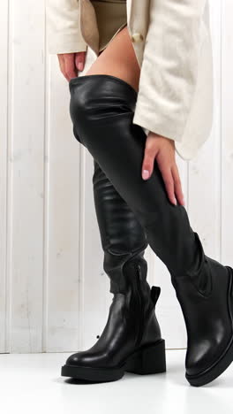 elegant very high black boots demonstration in studio. model in white mini skirt touching her footwear standing at the white wall. vertical video