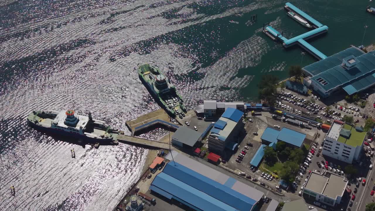 Aerial view of Victoria Jetty on Labuan Island, Malaysia, showing Ro-Ro ship, car carrier, and port buildings under sunlight reflecting on ocean waves. Concept of transport or maritime footage