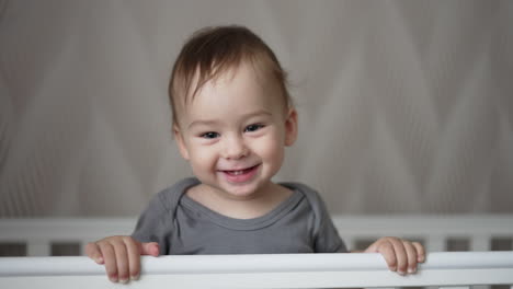 adorably smiling baby boy and showing his two lower teeth. sweet little toddler indoors in white baby cot. close up. blurred backdrop.