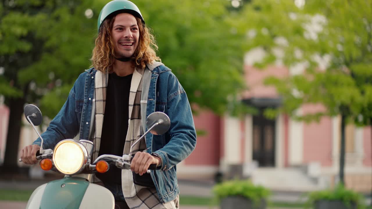 A guy with curly hair in a denim jacket and a green moped helmet drives up on his moped, meets his brunette girlfriend in a plaid shirt and they continue their journey together in a summer city