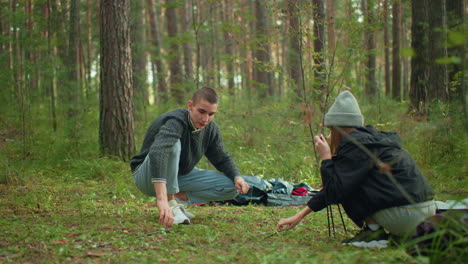 researcher dismantling tent pole during outdoor camp setup while woman bends to pick it up beside him in forest as he watches her warmly under soft natural daylight in peaceful forest
