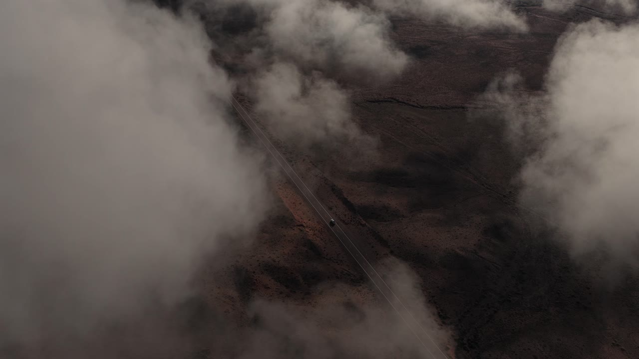Uma tomada de estabelecimento cinematográfica de drone segue um carro solitário dirigindo por uma estrada deserta remota. Névoa ascendente e o sol da manhã iluminam a paisagem acidentada das badlands nesta cena de viagem épica.