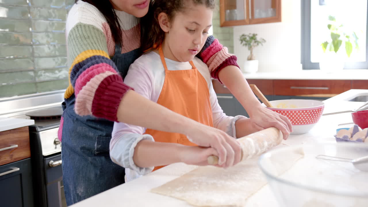 Happy biracial mother and daughter rolling out dough and smiling in sunny kitchen