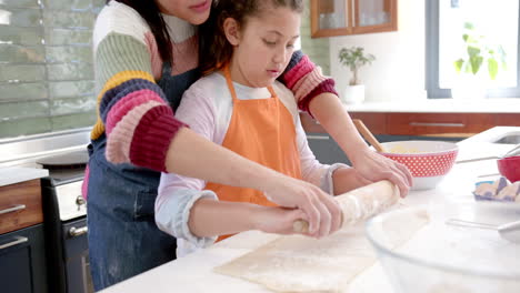 happy biracial mother and daughter rolling out dough and smiling in sunny kitchen