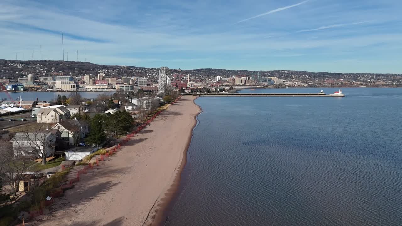Drone Flying over Beach next to houses and trees. Brown sand and blue water. Bridge and harbor in the back. Park Point in Duluth, Minnesota. Lake Superior