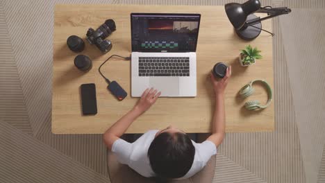 top view of asian woman editor drinking coffee while sitting in the workspace using a laptop next to the camera editing the video at home