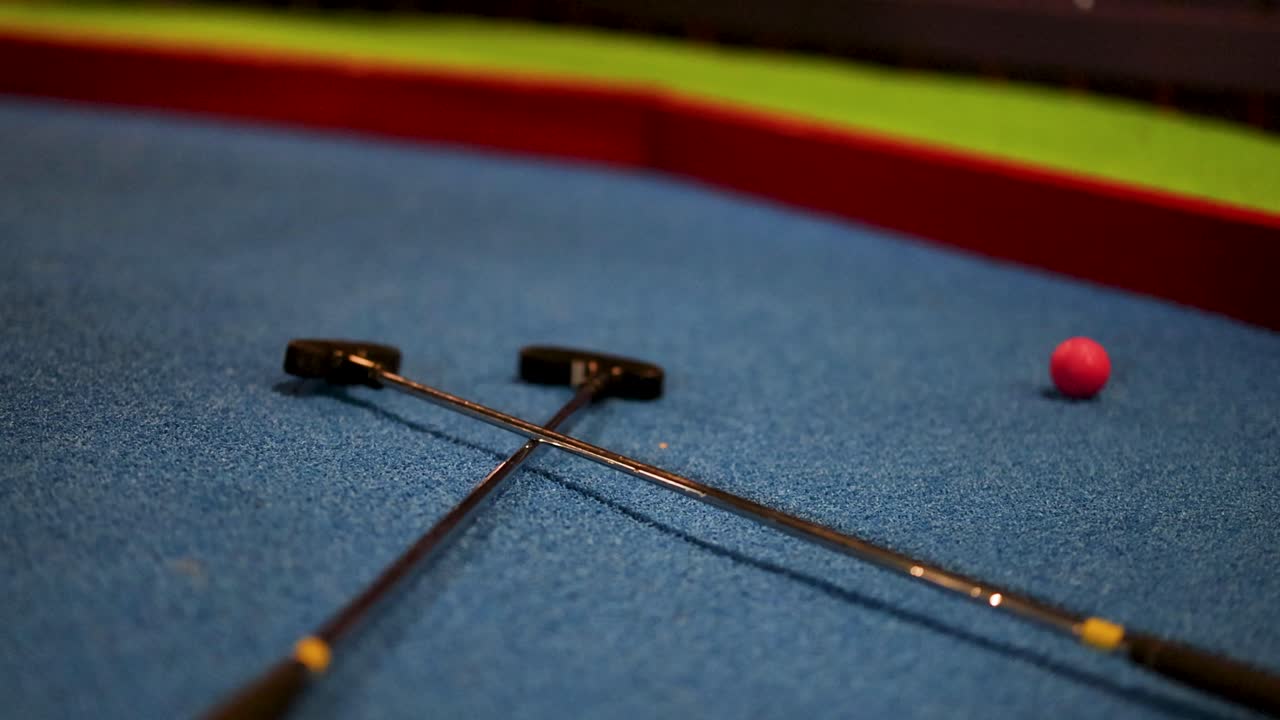 A pink golf ball bounces across a blue miniature golf course, passing two crossed putters under dim lighting
