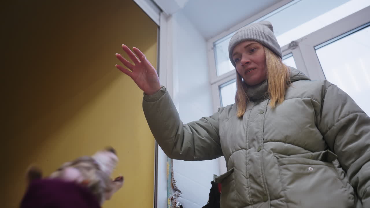 Woman in green winter jacket and gray beanie standing indoors near door and yellow wall, holding hand out with focused expression, possibly interacting with pet or preparing to give treat or command