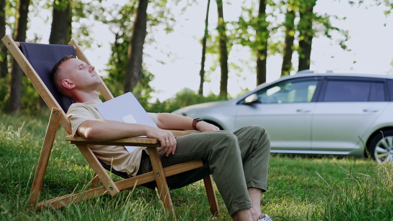 Freelancer sleeps in a folding chair with face covered with laptop. Tired man having a break from work in the nature.