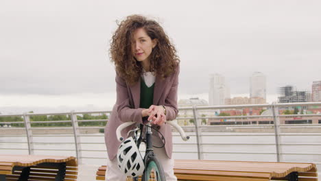 beautiful curly woman in formal clothes looking and smiling at the camera while sitting on a bicyle on the city bridge