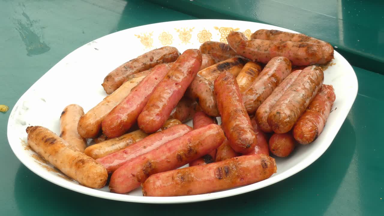 A plate of small barbecued mini cocktail sausages. A hand comes into frame and picks up on sausage.