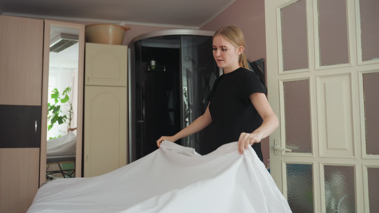 Woman dressed in black spreads white bed sheet over mattress in softly lit interior room with brown wardrobe behind her, captured in mid-motion as fabric floats above bed, evoking sense of routine