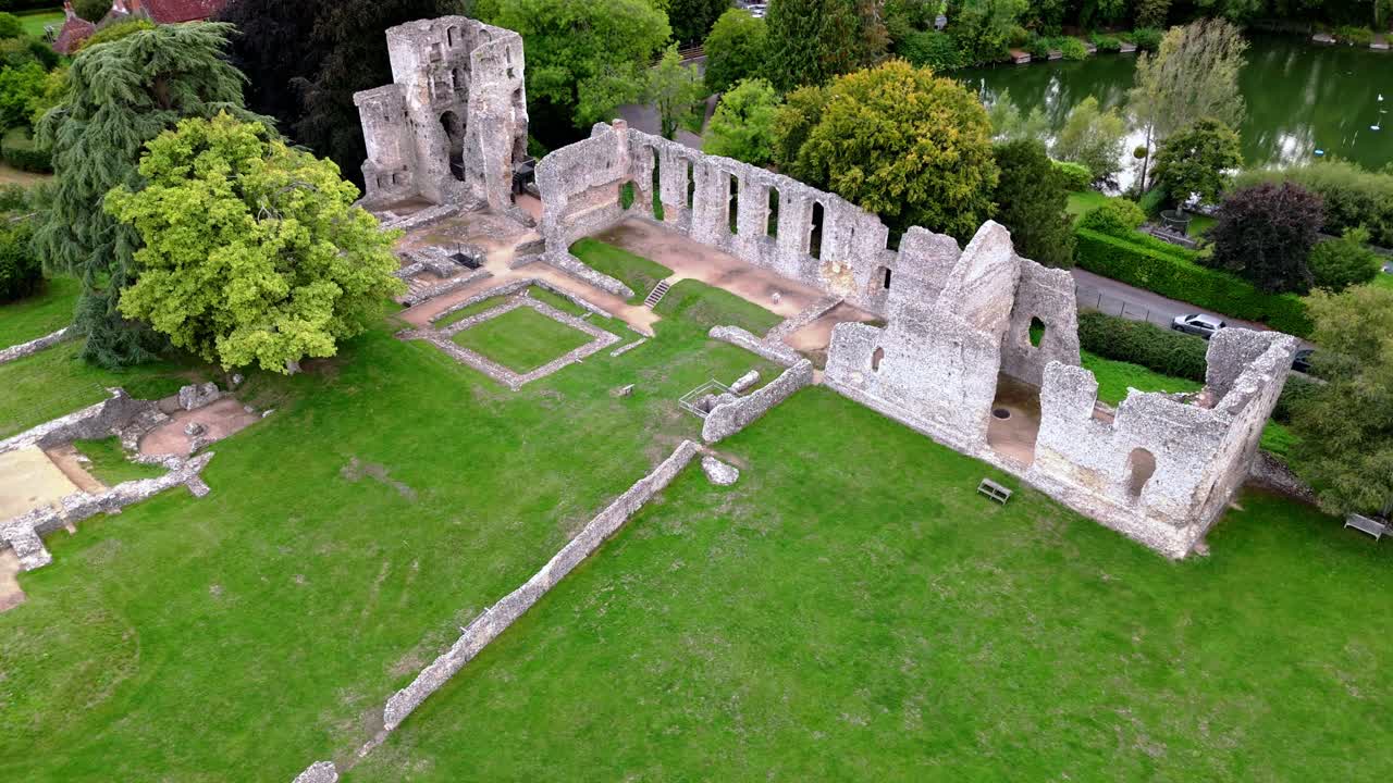 Low-angle aerial drone pans downward over Bishop’s Waltham Palace ruins and pond, capturing historic walls, autumn trees, and warm golden sunset light across the tranquil village scene