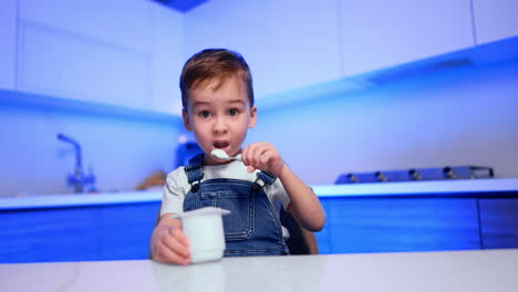 beautiful toddler baby wearing jeans romper eats yogurt at the table. low angle view at the kid having a healthy snack himself.