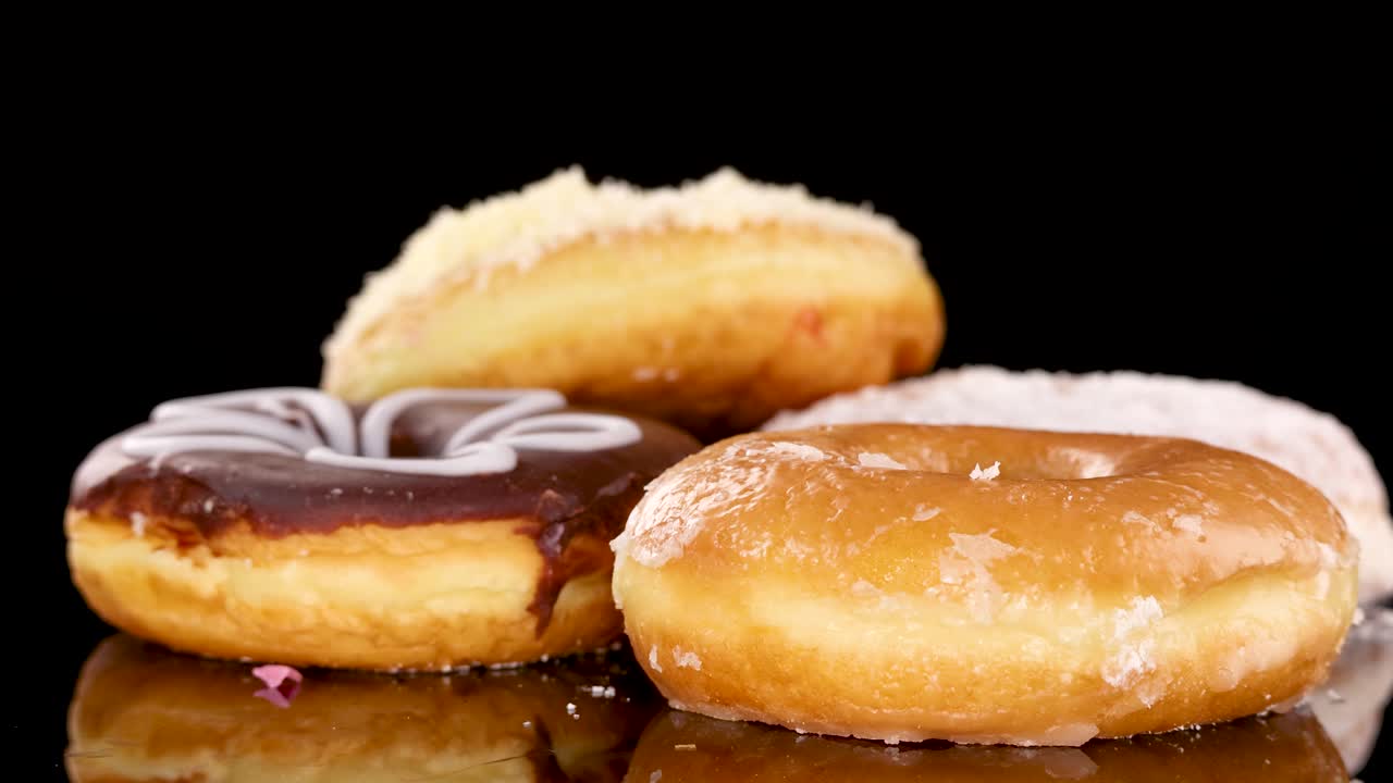 A chocolate-glazed, white-iced donut ring drops onto a stack of assorted donuts on a reflective surface, under bright studio lighting with a black background
