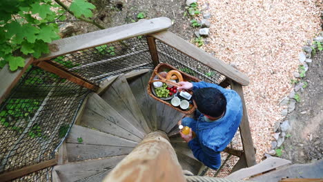 Static overhead FHD shot of a man in a denim jacket and jeans climbing a spiral wooden staircase carrying a basket with breakfast
